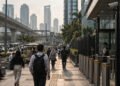 Pejalan kaki di trotoar kawasan bisnis Jakarta dengan gedung tinggi, lalu lintas padat, pagar, CCTV, dan area keamanan.
